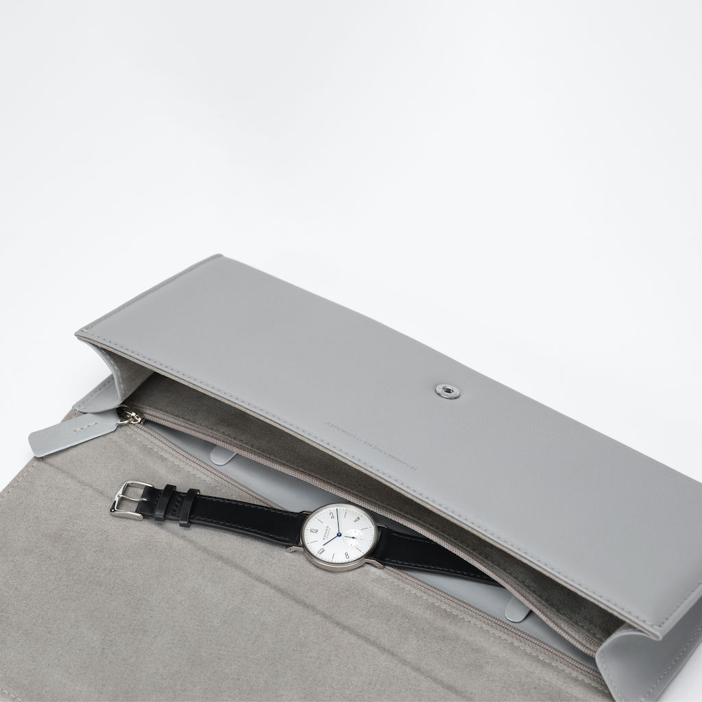 Clutch NOMOS, fabricado en cuero de vaca resistente a la luz y curtido sin cromo, forrado. Dimensiones: 30 x 12,5 cm. Cierre: botón de presión sujeto mediante costura. Compartimiento individual para reloj, con cremallera.